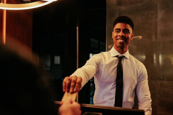 A young African American receptionist is servicing a hotel guest at the front desk by handing his room keys over the counter.